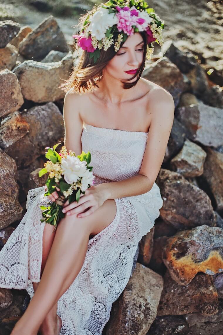 bride sitting on rocks on beach holding flowers wearing flower crown 