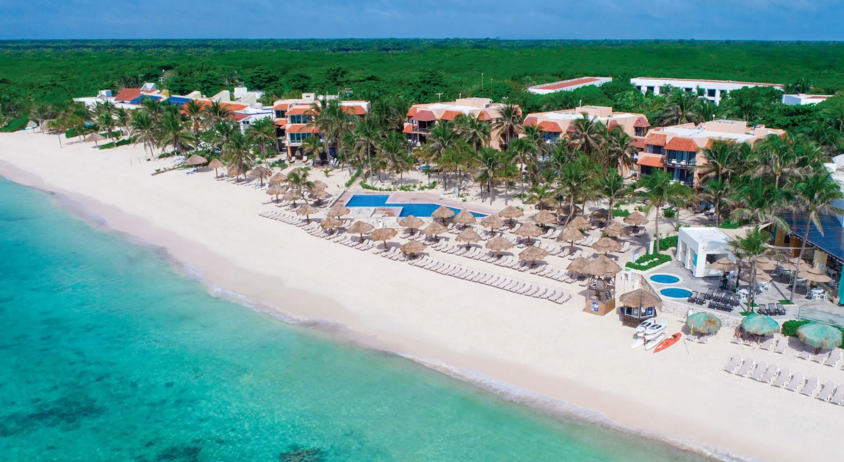Aerial panoramic view of a beachfront resort with white sand, palm trees, and turquoise water