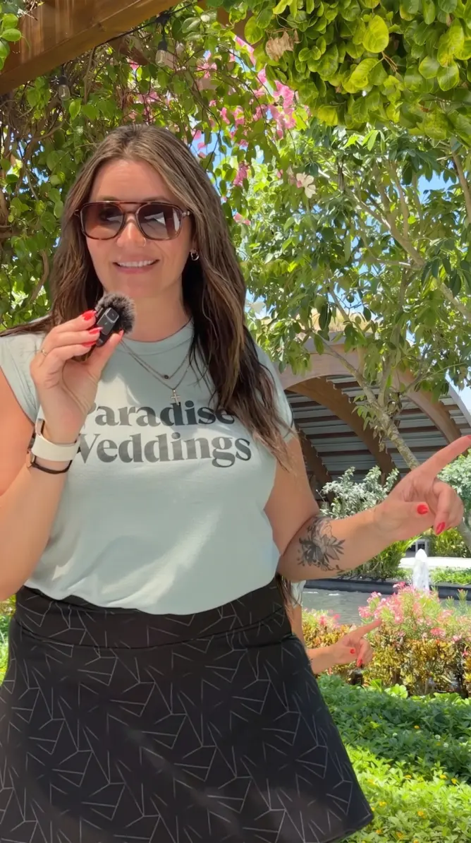 Paradise Weddings planner Lindsay speaking into a microphone outdoors under a floral pergola