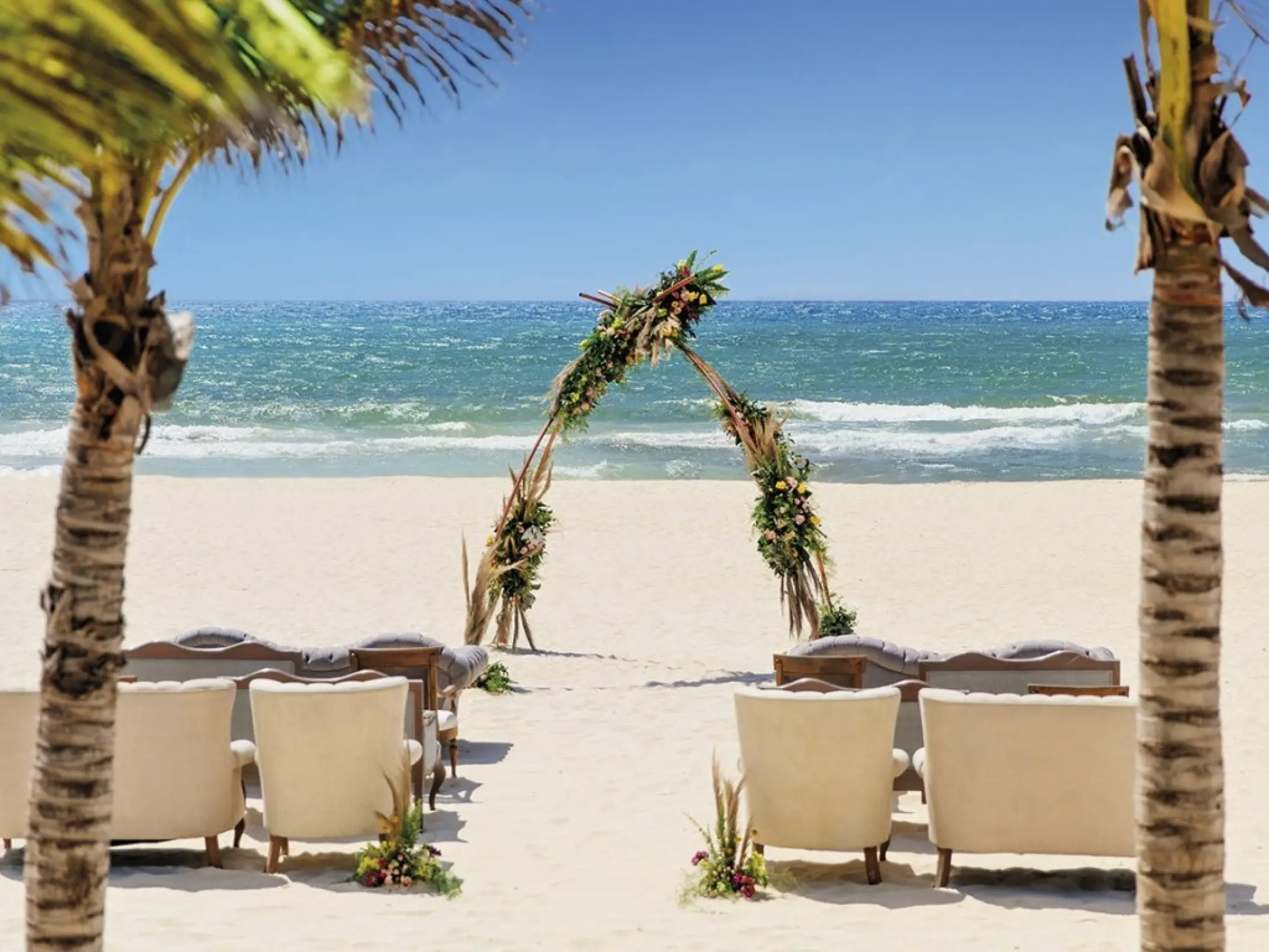Beachfront wedding ceremony setup with triangular floral arch and white armchairs at Allegro Playacar, Playa Del Carmen