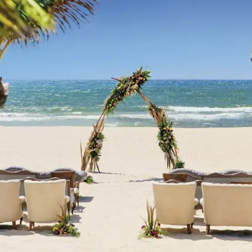 Beachfront wedding ceremony setup with triangular floral arch and white armchairs at Allegro Playacar, Playa Del Carmen