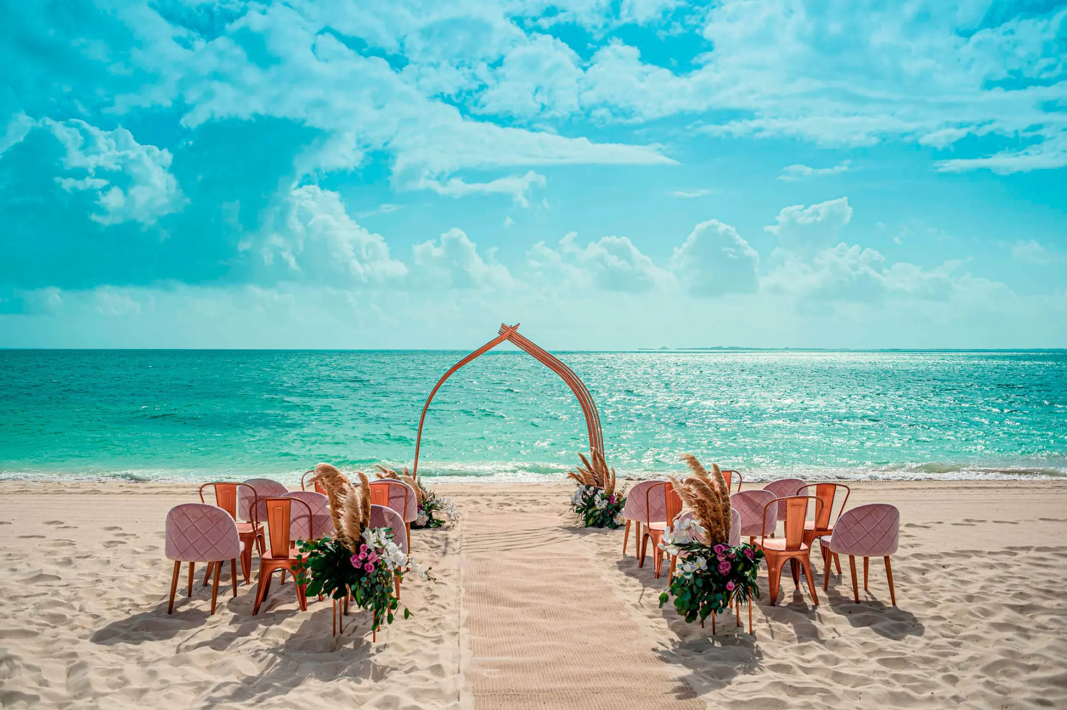 Beachfront wedding ceremony setup with a floral arch and pink chairs at Atelier Playa Mujeres, Cancun