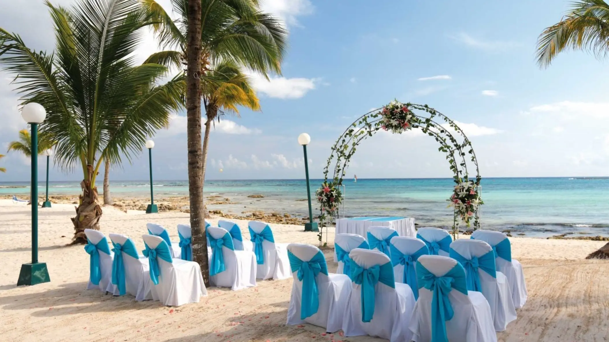 Beachfront wedding ceremony setup with blue chair sashes and floral arch at Barcelo Maya Riviera, Riviera Maya