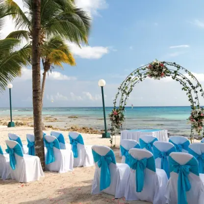 Beachfront wedding ceremony setup with blue chair sashes and floral arch at Barcelo Maya Riviera, Riviera Maya
