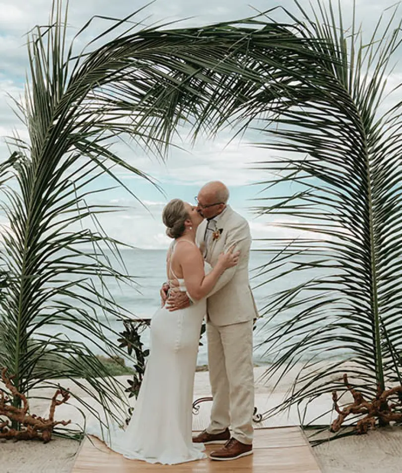 Carrie & David kissing under a palm frond archway during their beach ceremony