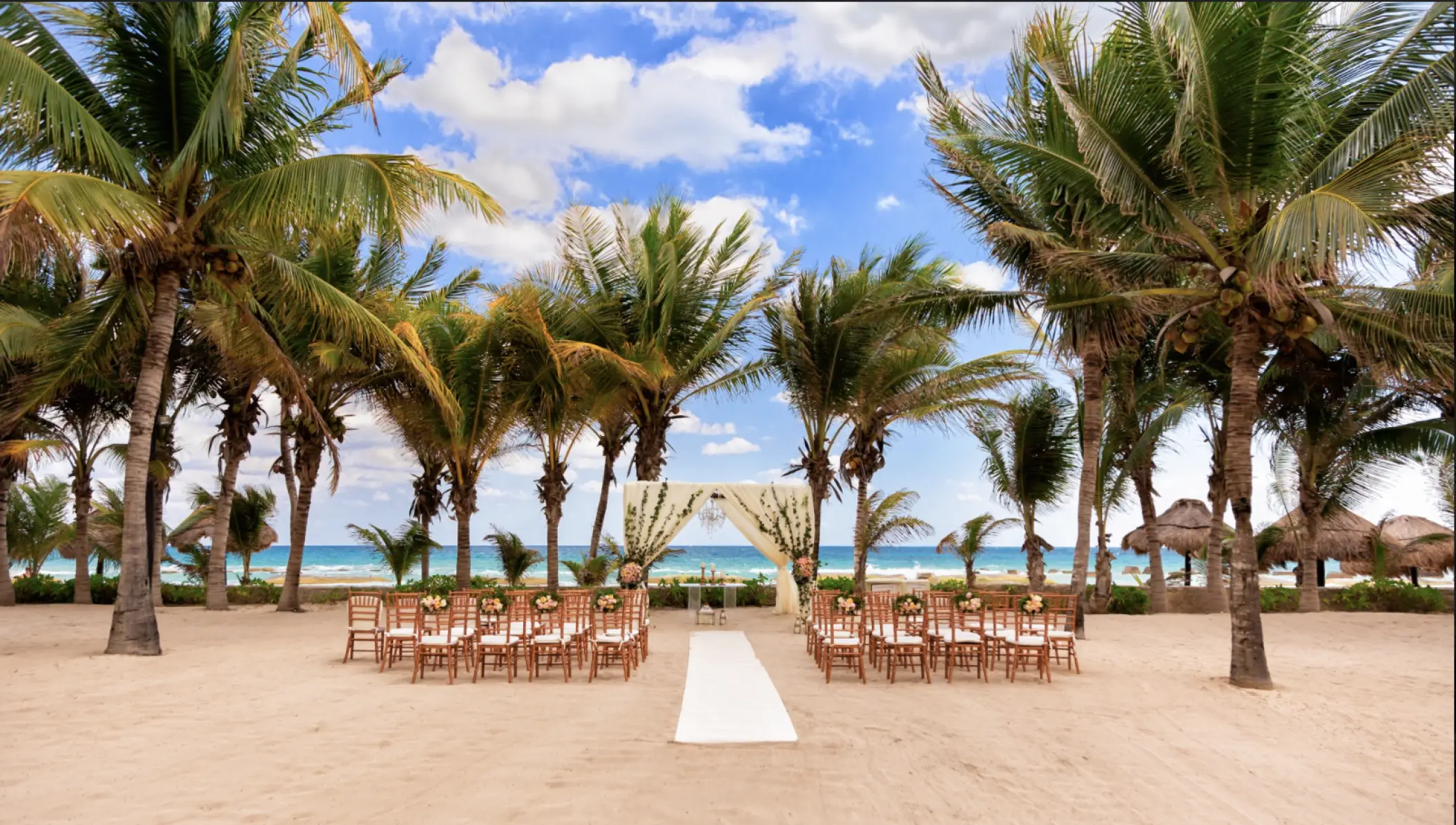 Beachfront wedding ceremony setup with wooden chairs and palm trees at El Dorado Royale, Riviera Maya