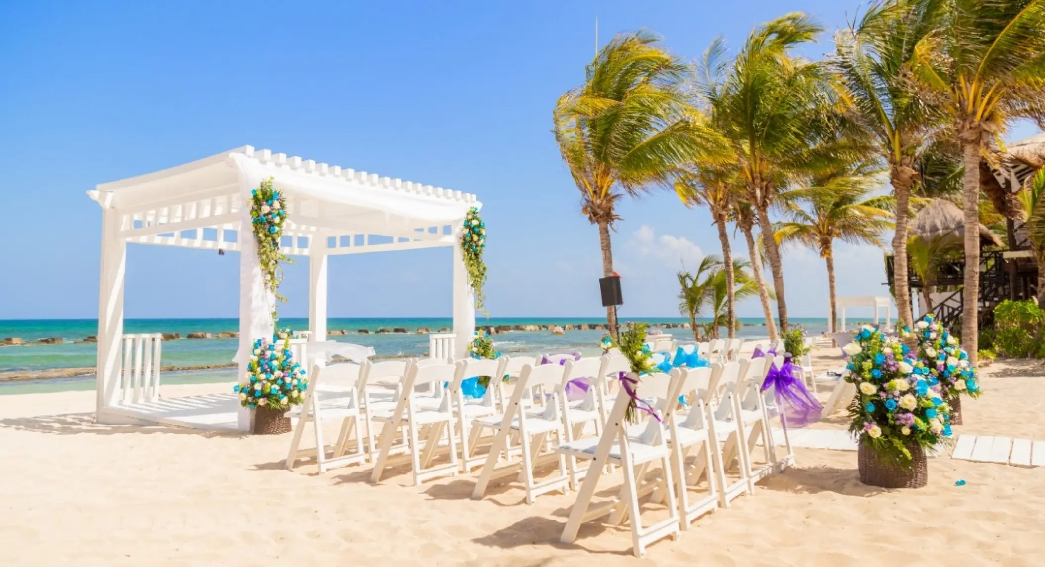 Beachfront wedding ceremony with white pergola and blue flower arrangements at El Dorado Royale, Riviera Maya