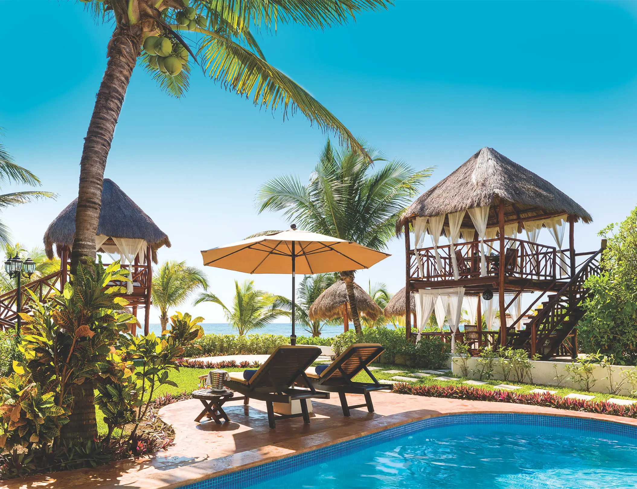 Poolside lounge chairs under a parasol with thatched cabanas at El Dorado Seaside Palms, Riviera Maya