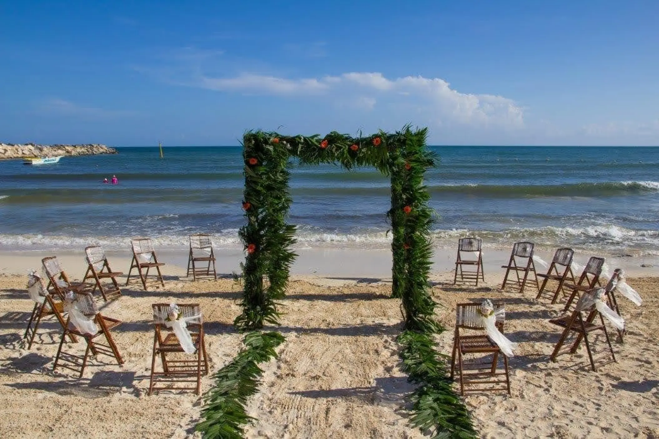 Beachfront wedding ceremony with a greenery arch and wooden chairs at Dreams Jade, Riviera Maya