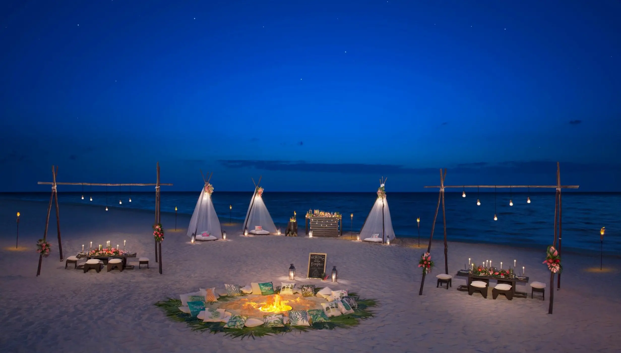 Beachfront wedding reception with fire pit and lit tipi tents at night at Dreams Riviera Cancun, Riviera Maya