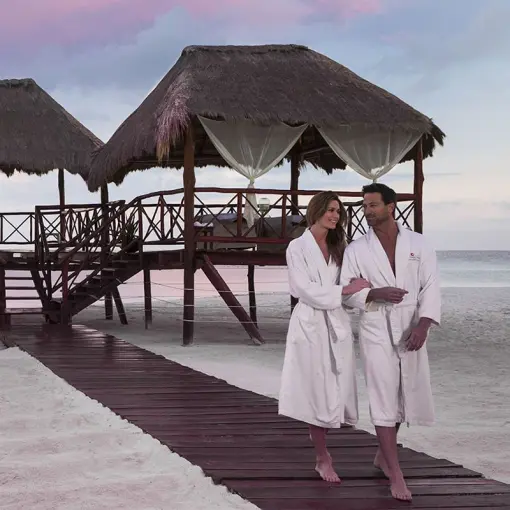 Couple in white robes walking on a wooden path by beach spa palapas at El Dorado Maroma, Riviera Maya
