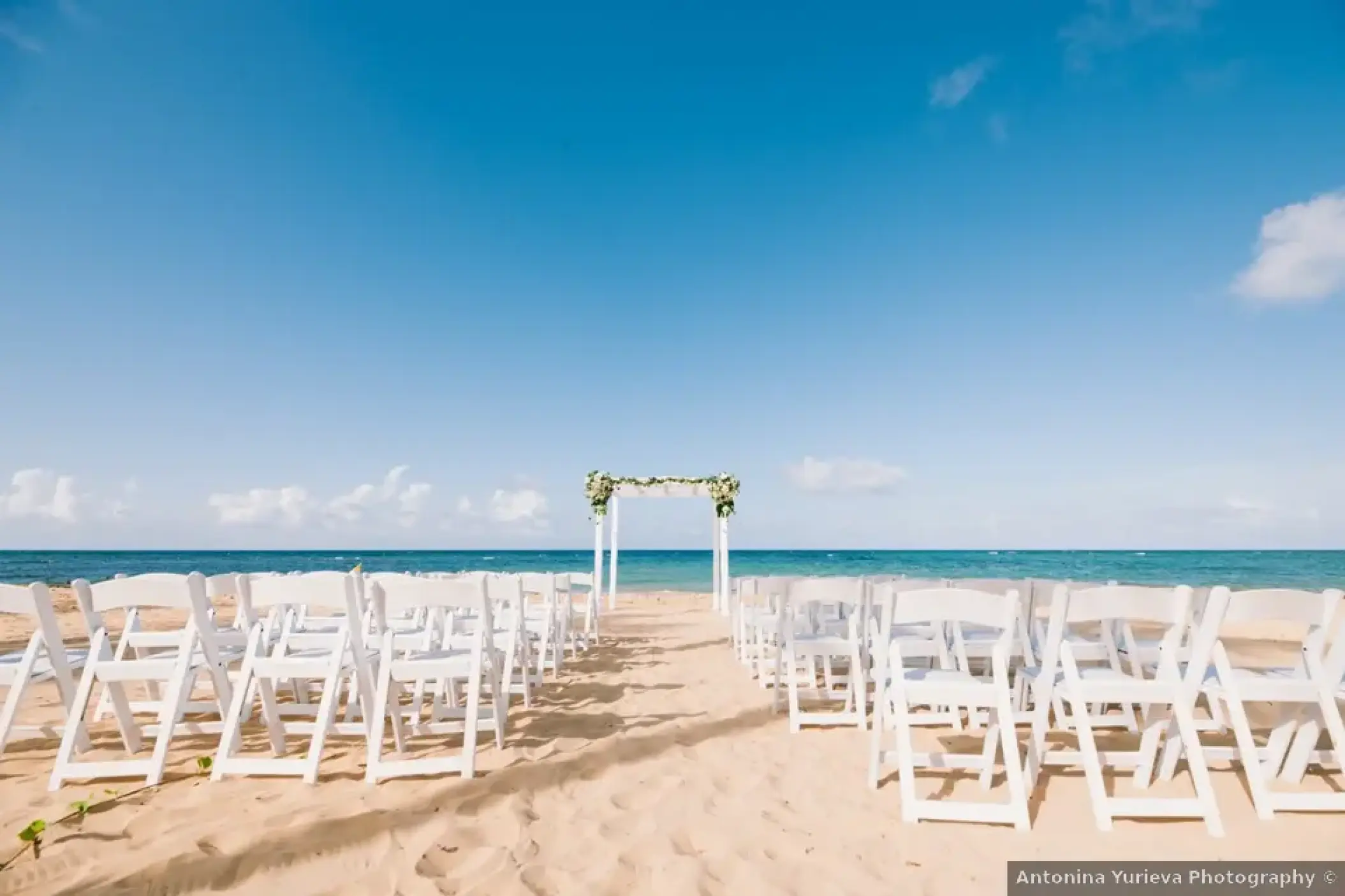 Beachfront wedding ceremony with white chairs and floral arch at Excellence El Carmen, Punta Cana