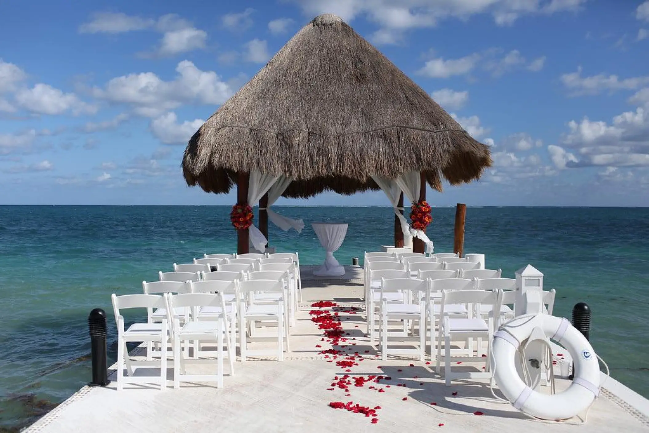 Pier wedding ceremony under a palapa with red flower petals and white chairs at Excellence Riviera Cancun