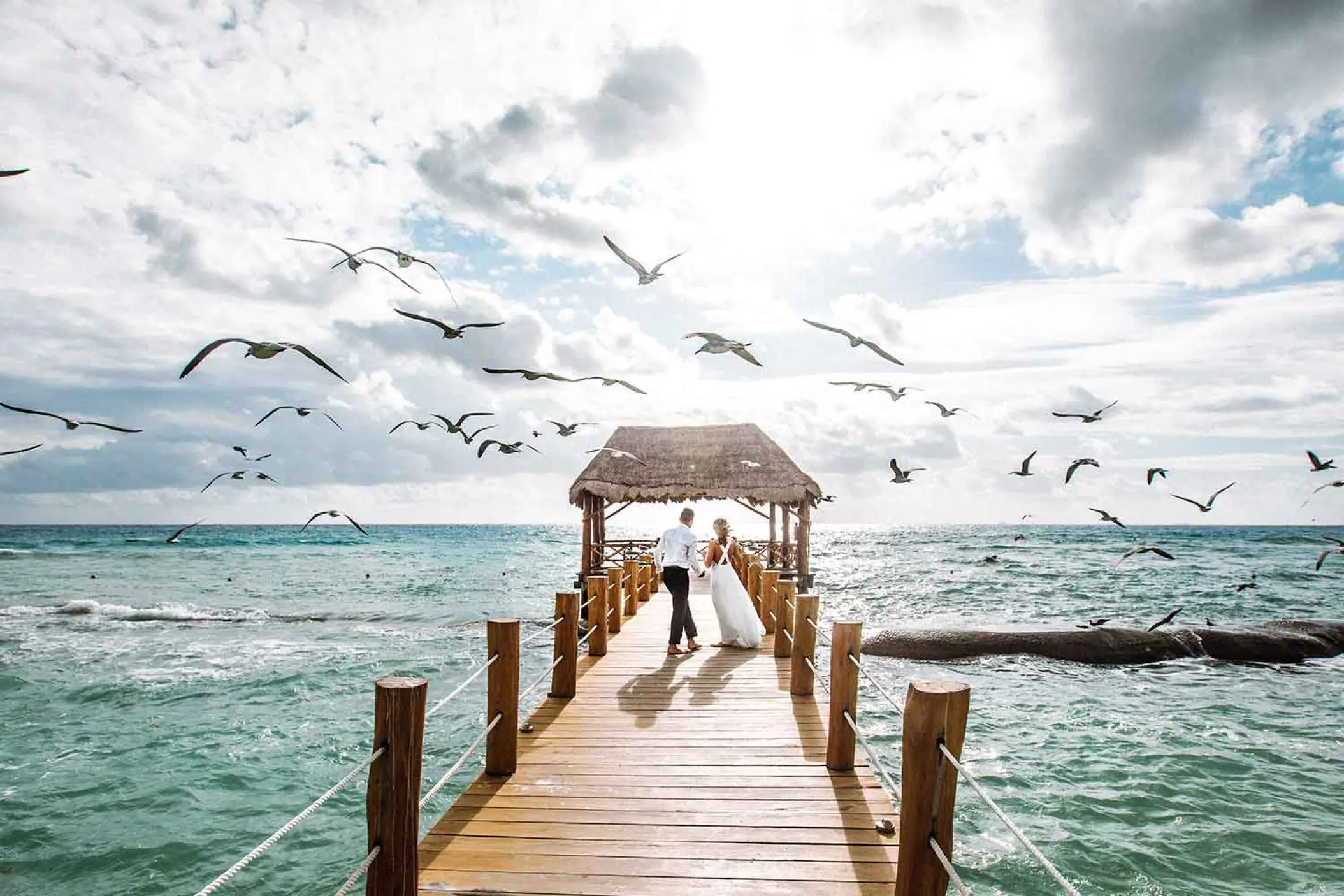 Bride and groom standing on the pier at The Fives Beach Hotel, Playa Del Carmen as birds fly overhead