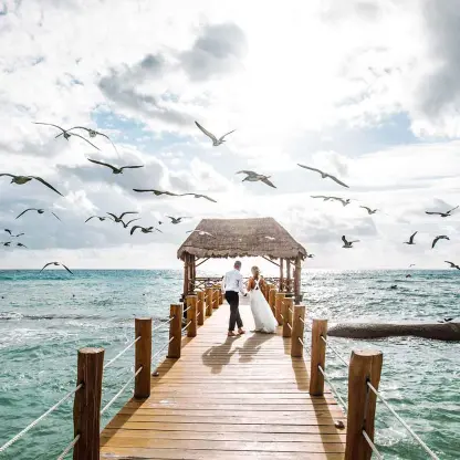 Bride and groom standing on the pier at The Fives Beach Hotel, Playa Del Carmen as birds fly overhead