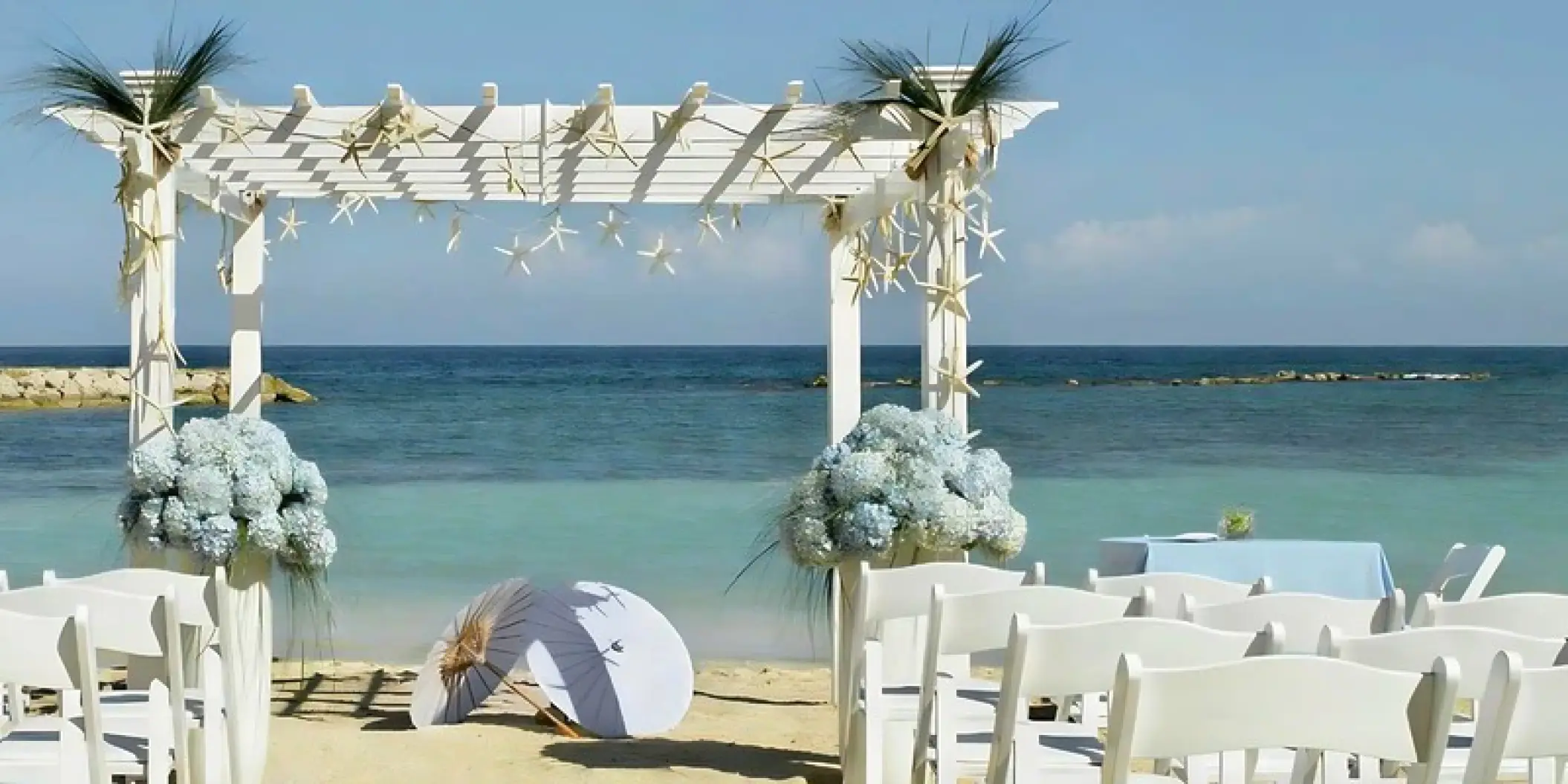 Beachfront wedding ceremony setup with white pergola and blue hydrangeas at Grand Palladium Jamaica, Ocho Rios