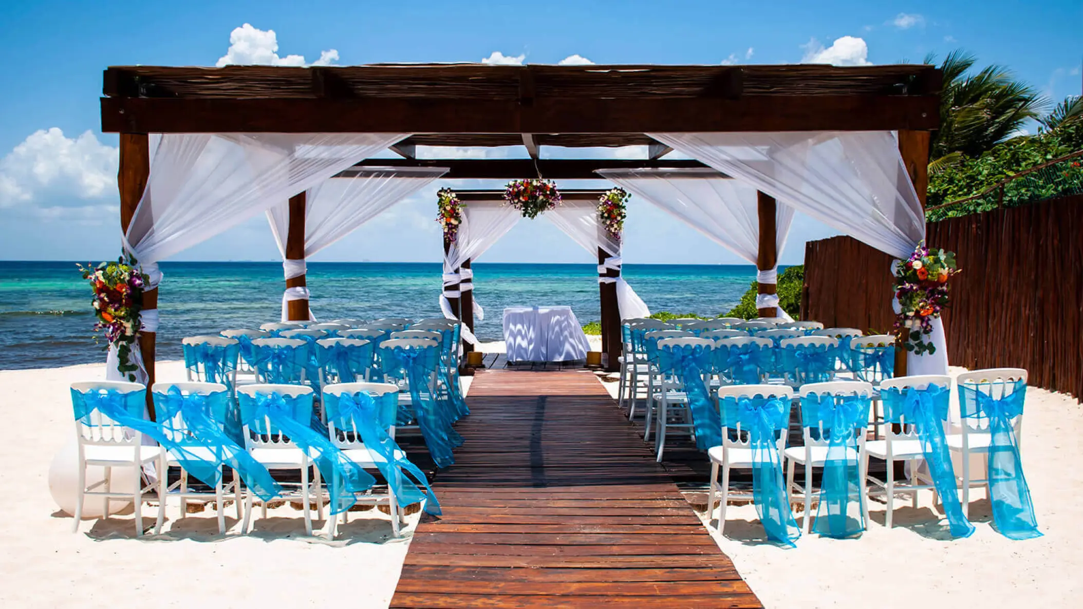 Beachfront wedding ceremony with blue-draped chairs under a wooden gazebo at Grand Riviera Princess, Playa Del Carmen