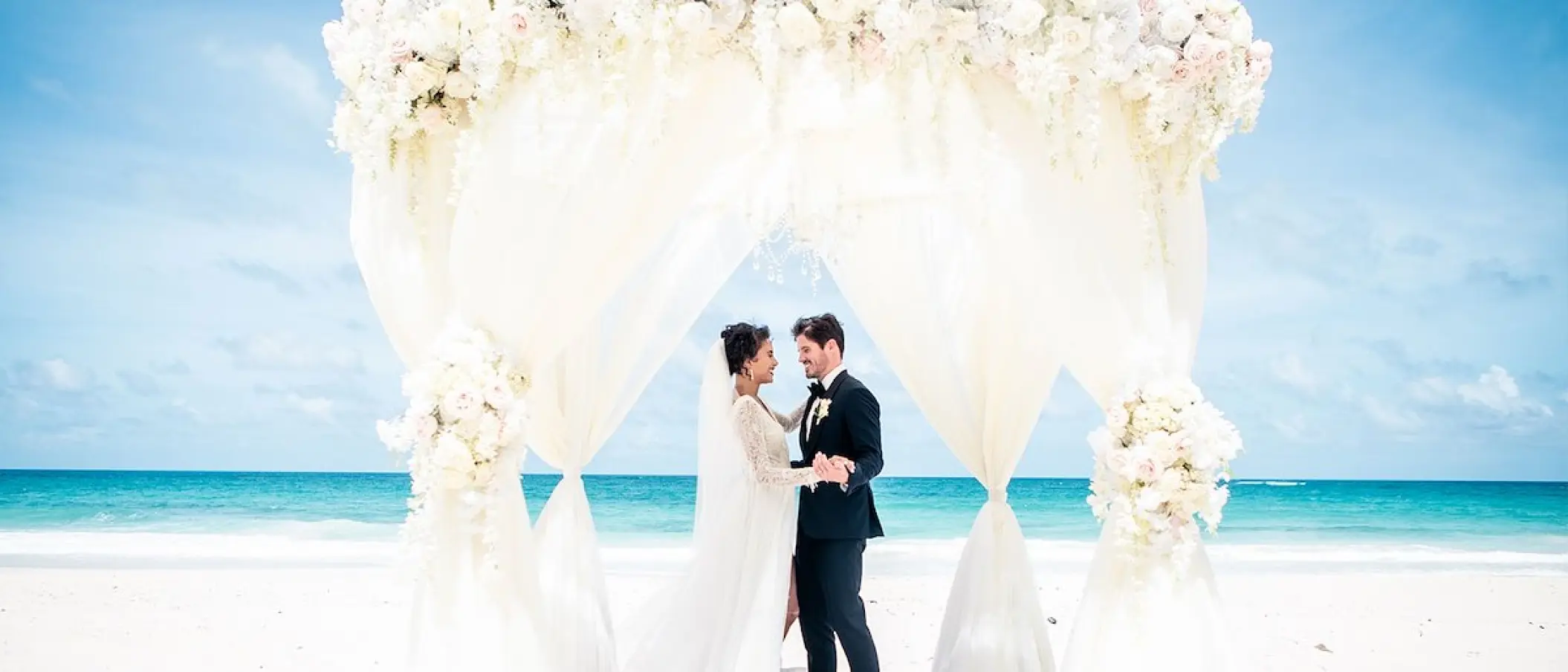 Bride and groom under a white draped floral arch for a beach ceremony at Hard Rock Cancun