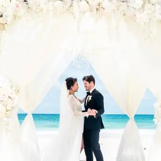 Bride and groom under a white draped floral arch for a beach ceremony at Hard Rock Cancun
