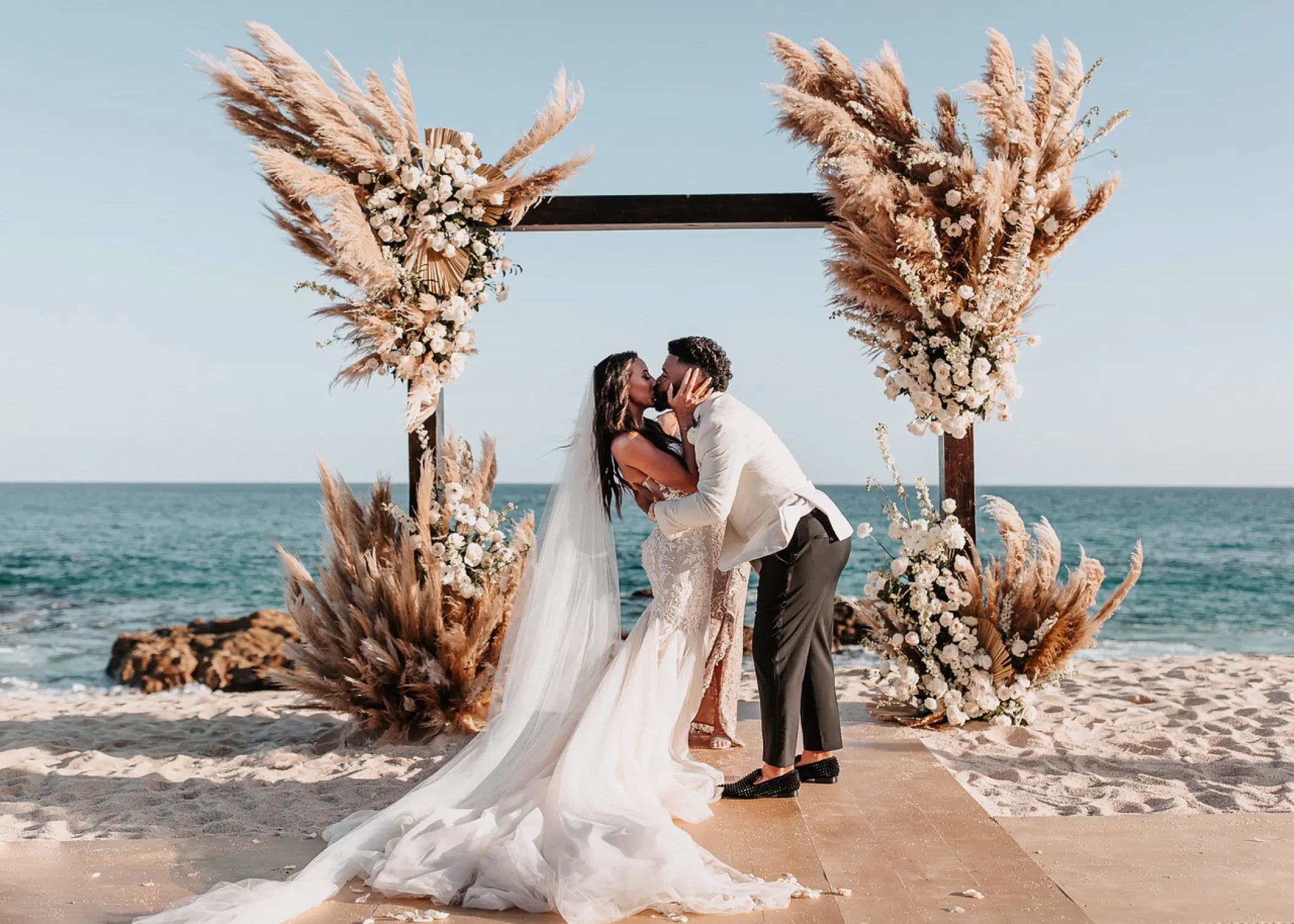 The bride and groom kissing under a pampas grass arch for a beach ceremony at Hilton Los Cabos, Cabo San Lucas