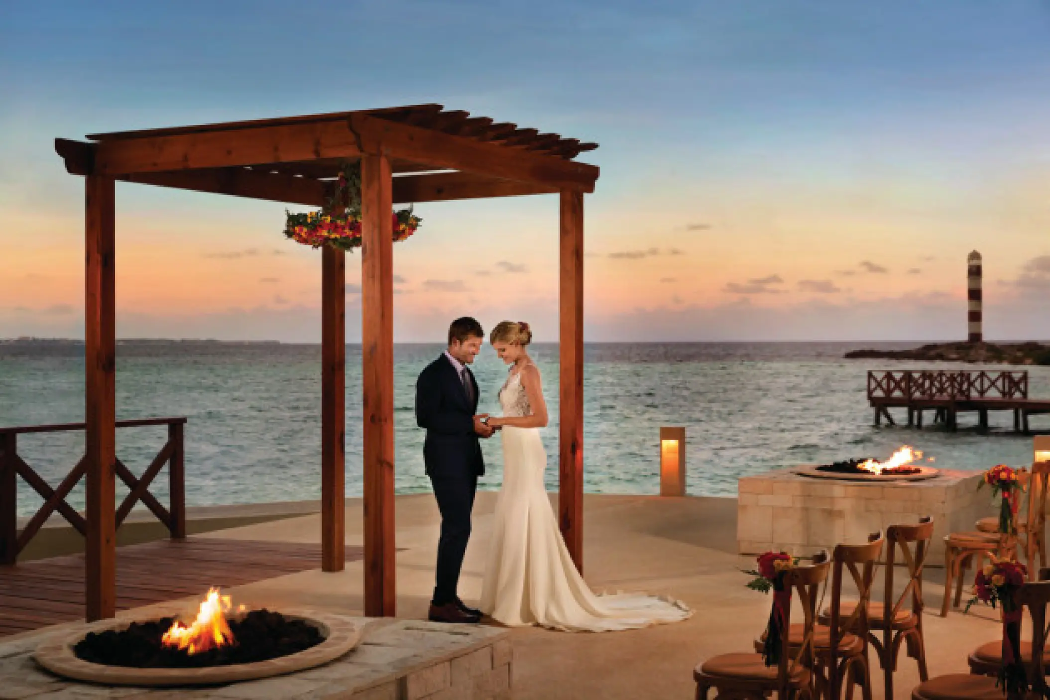 Bride and groom under a wooden gazebo at Hyatt Ziva Cancun, featuring fire pits and a lighthouse at sunset