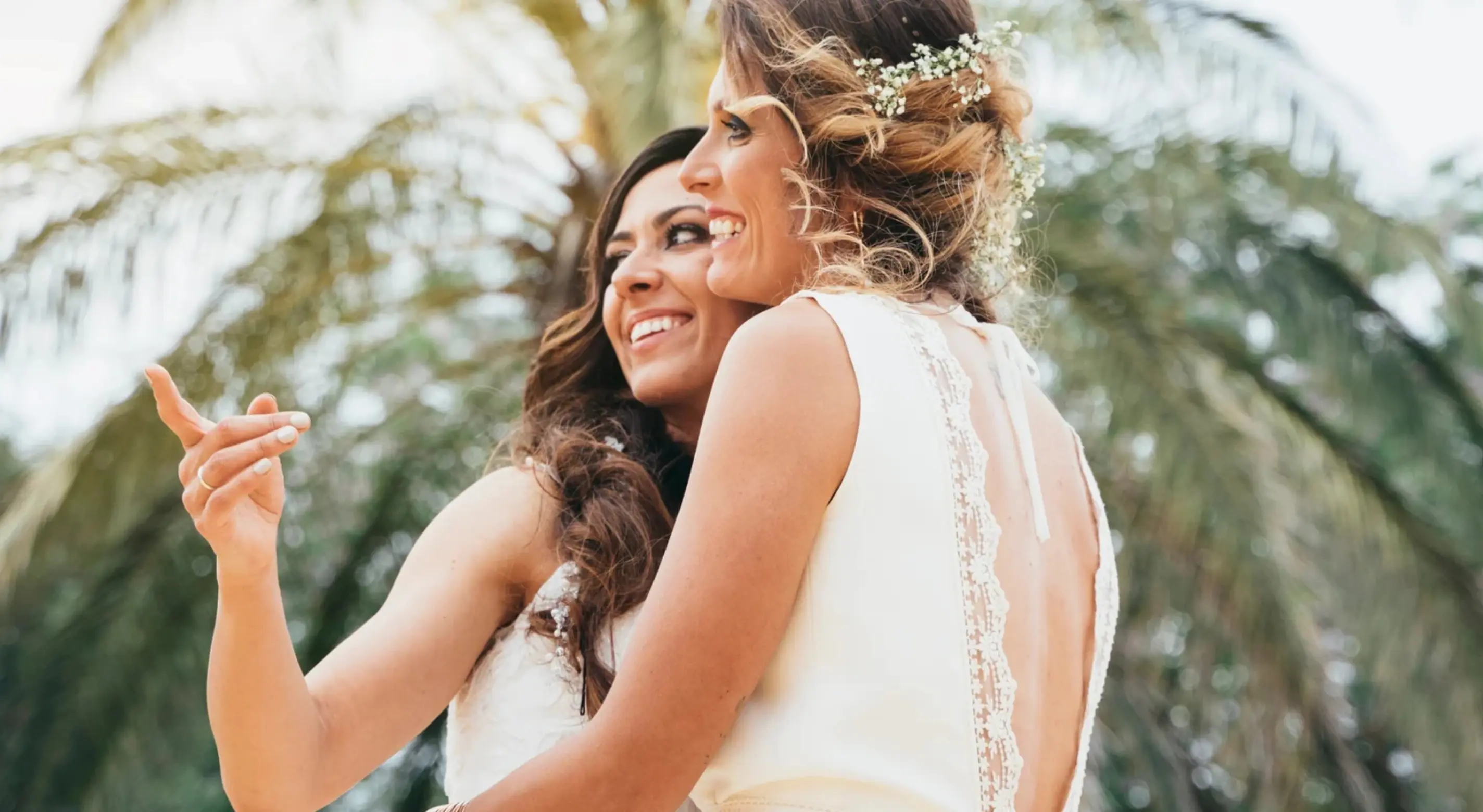 Two brides smiling and hugging under palm trees during a tropical wedding