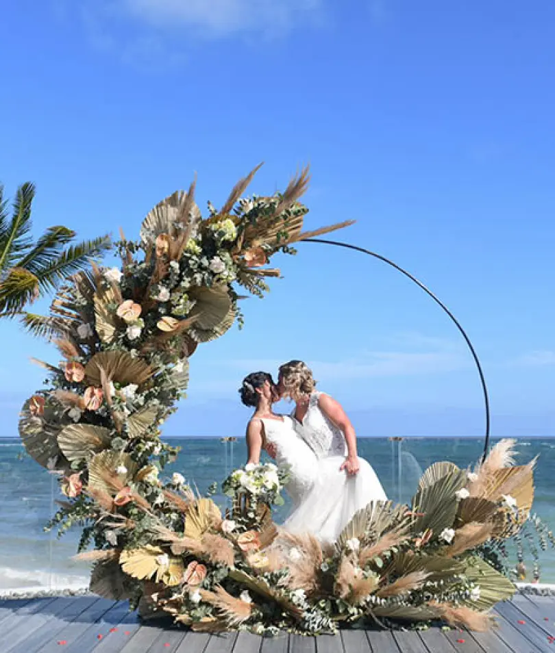 Stephanie & Kasey kissing inside a circular floral wedding arch on a terrace by the ocean