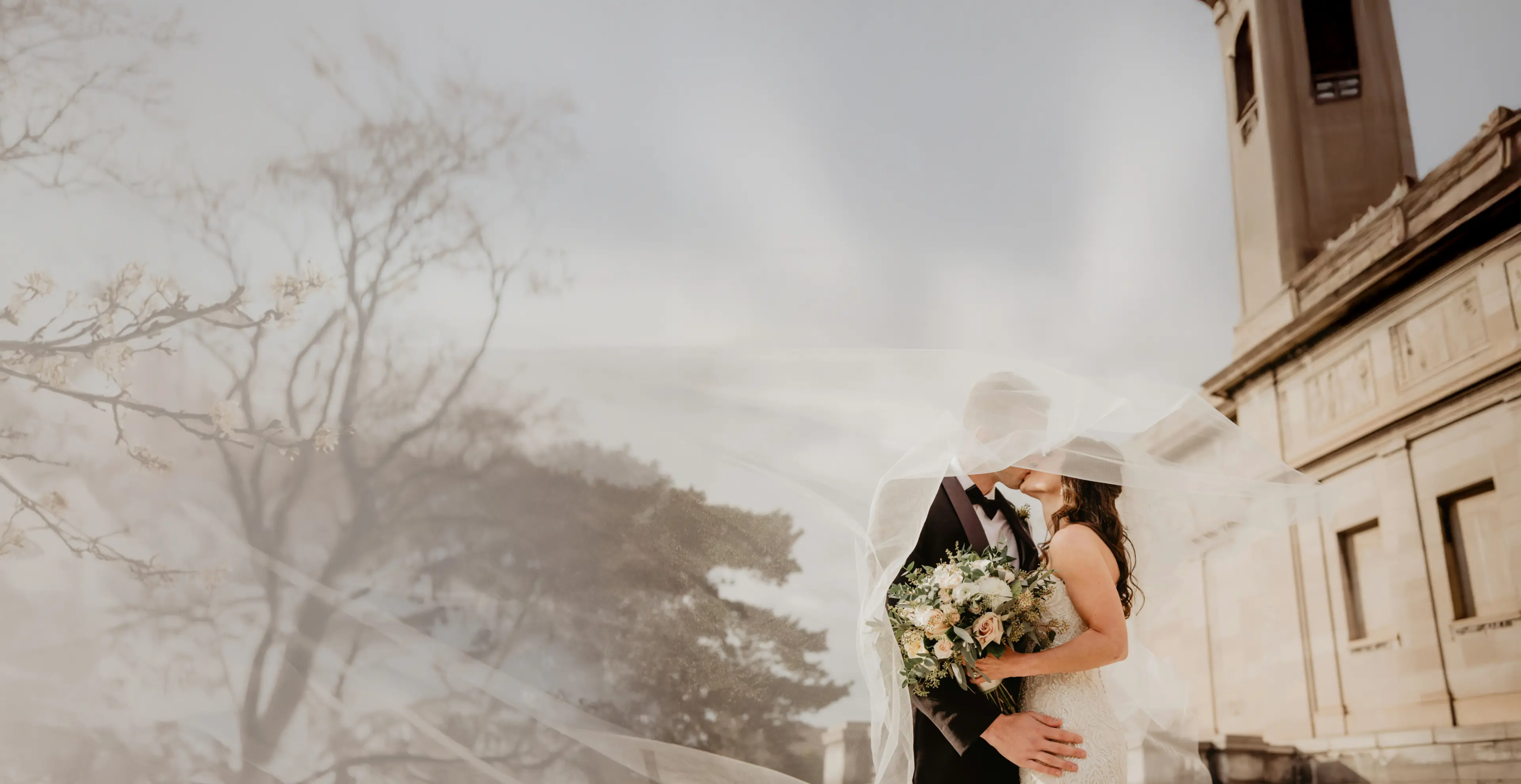The bride and groom sharing a kiss under a veil outside a stone chapel