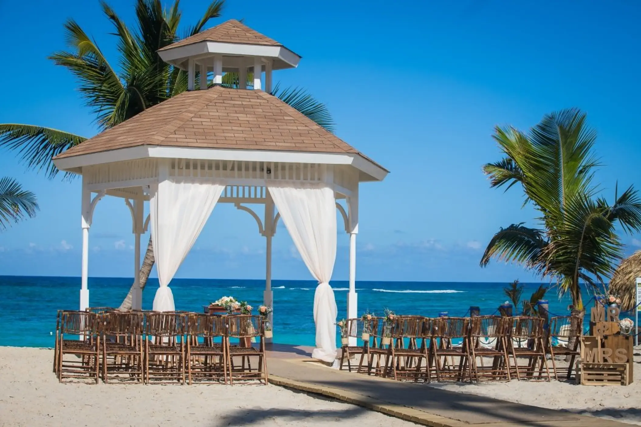 Beachfront wedding ceremony gazebo with white drapes and wooden chairs at Majestic Mirage, Punta Cana