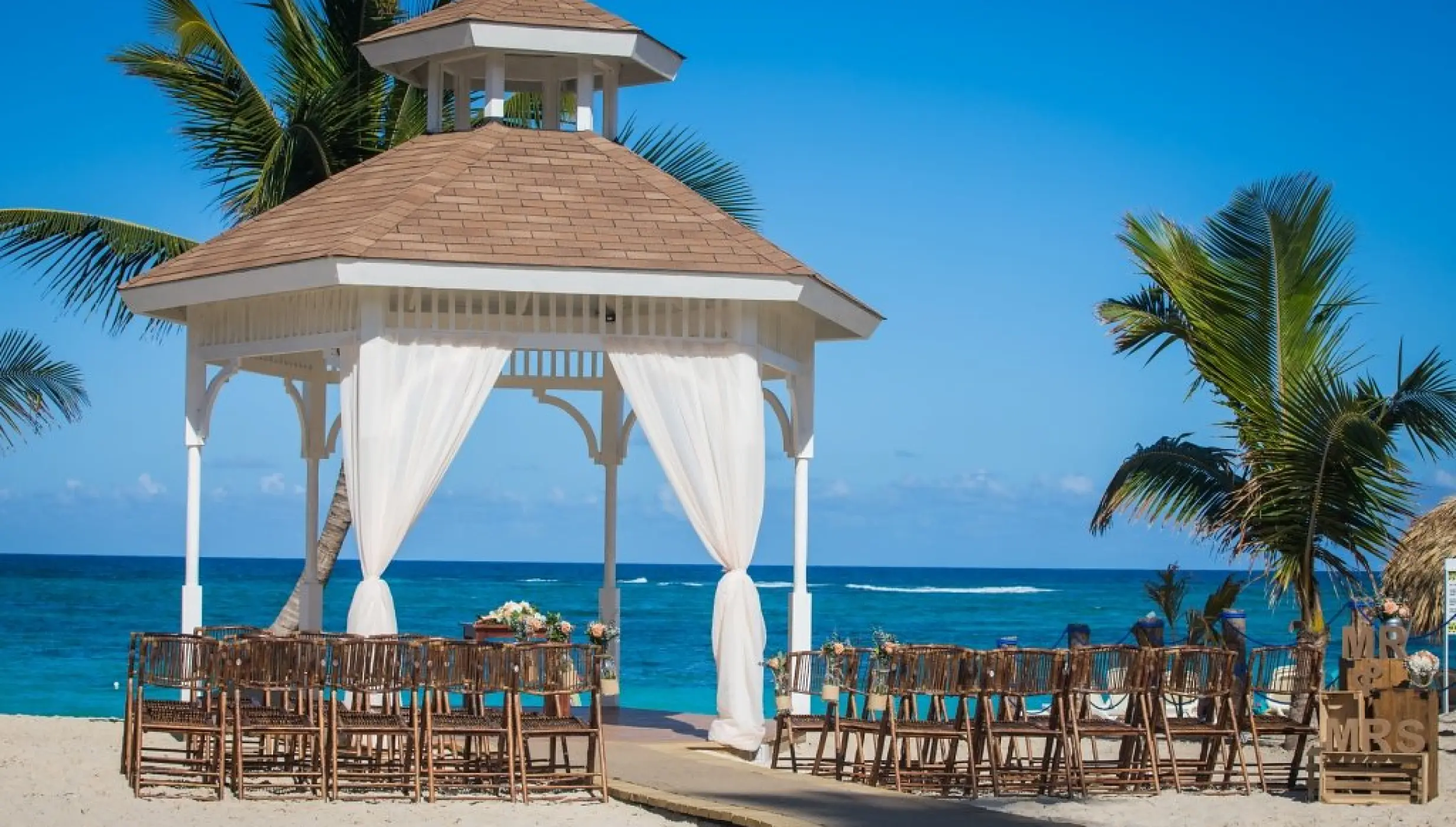 Beachfront wedding ceremony gazebo with white drapes and wooden chairs at Majestic Mirage, Punta Cana
