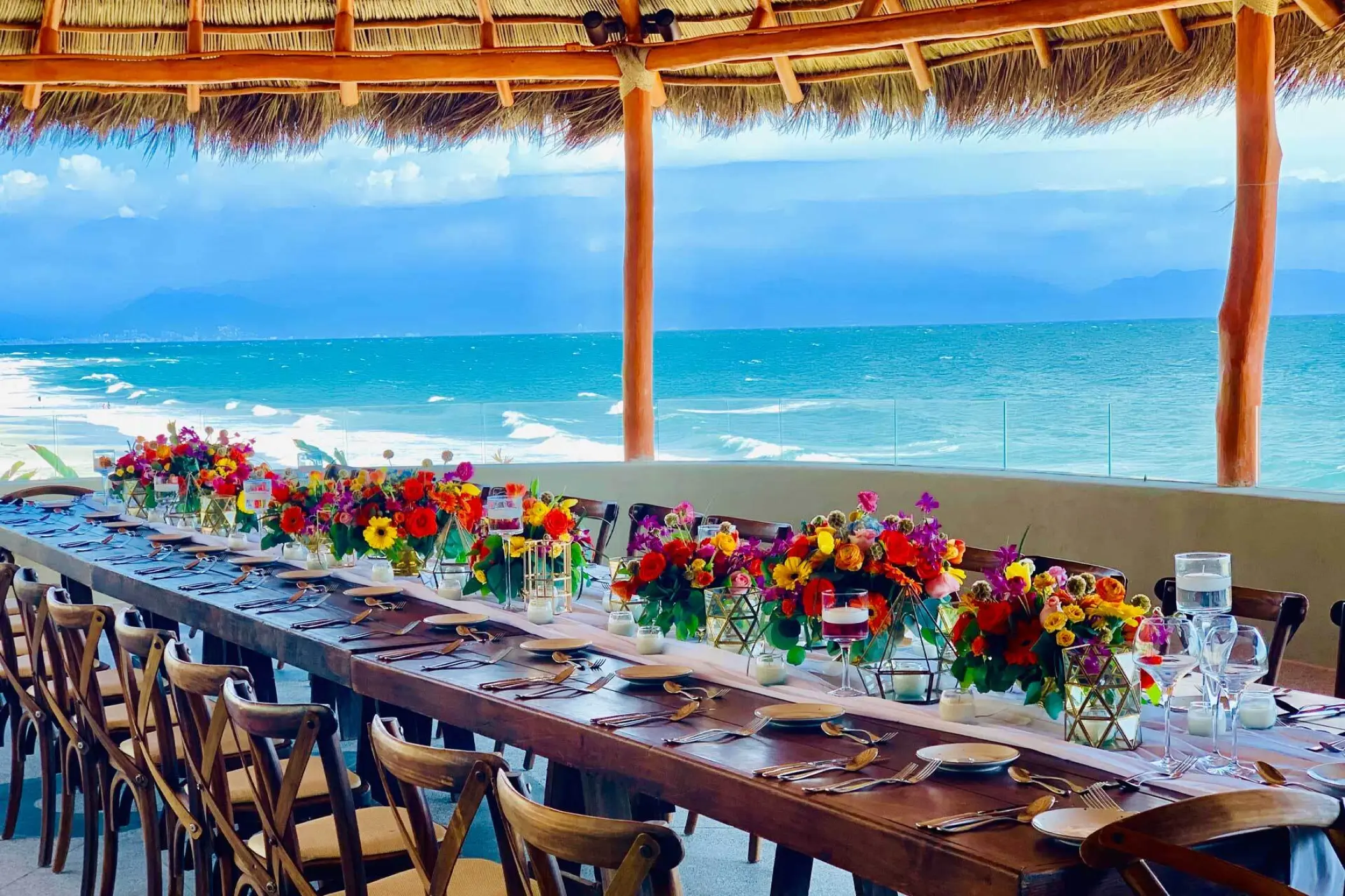 Long wooden reception table under a palapa with colorful floral centerpieces at Armony Marival, Puerto Vallarta