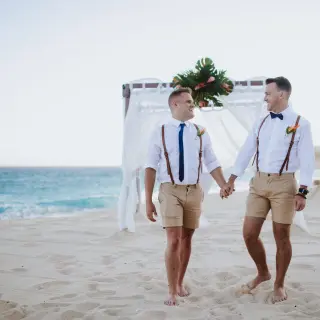Grooms holding hands on the sand during a beach ceremony at Marquis Los Cabos, Cabo San Lucas