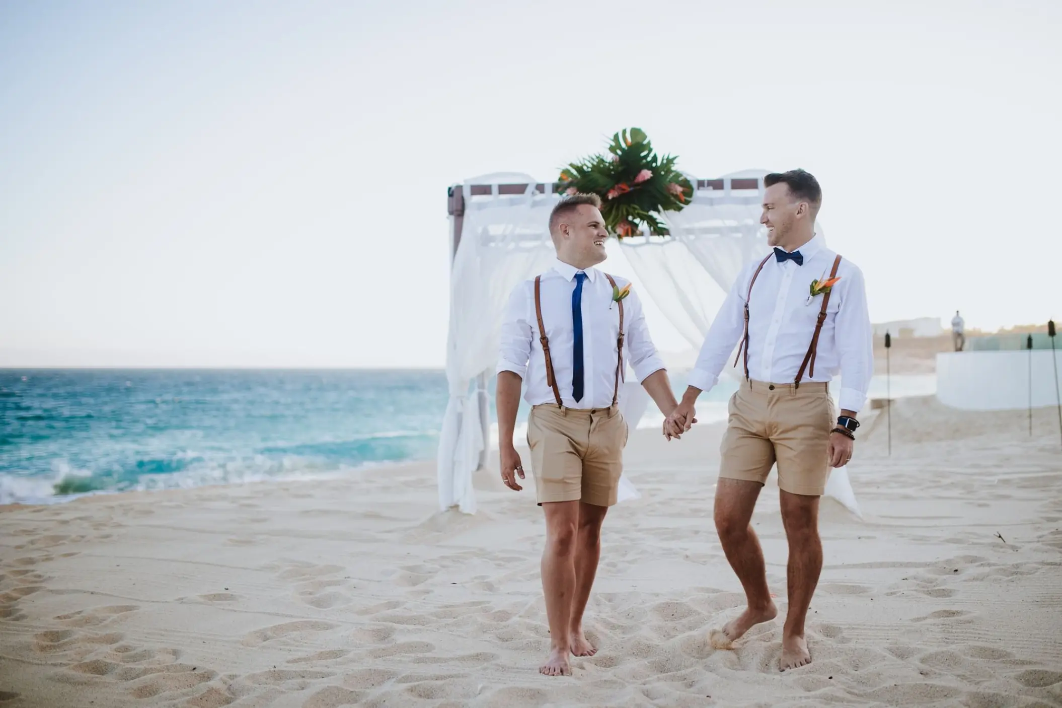 Grooms holding hands on the sand during a beach ceremony at Marquis Los Cabos, Cabo San Lucas