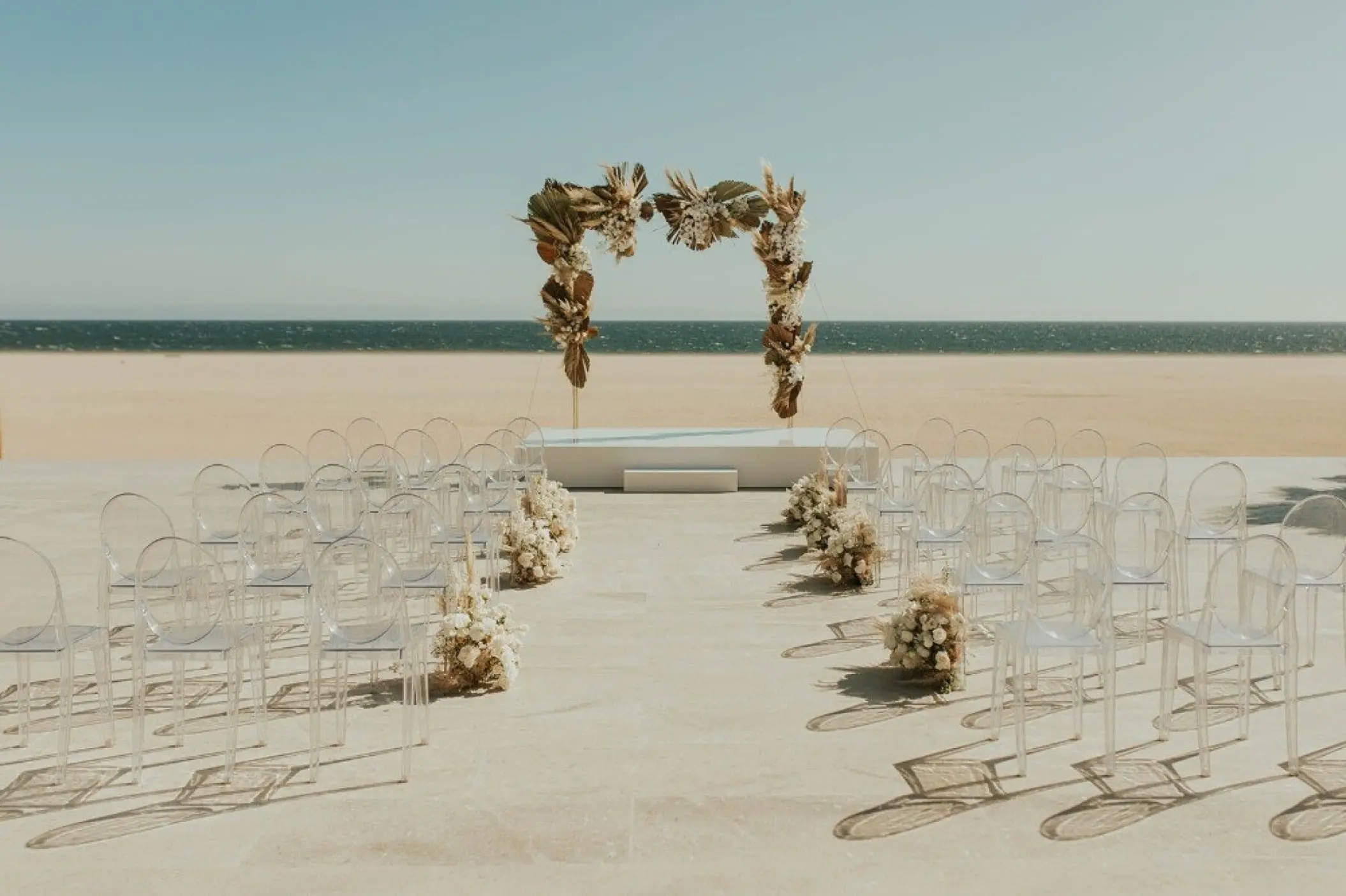 Beachfront wedding ceremony with clear chairs and a dried floral arch at Nobu Los Cabos, Cabo San Lucas