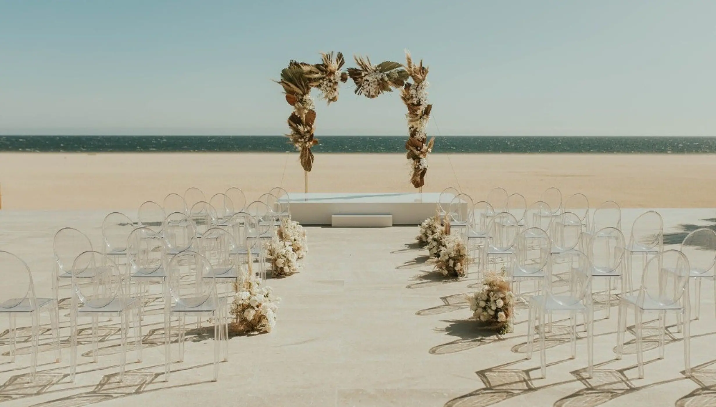 Beachfront wedding ceremony with clear chairs and a dried floral arch at Nobu Los Cabos, Cabo San Lucas