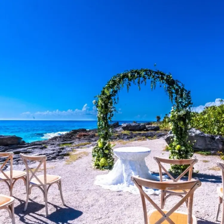 Rocky beachfront wedding ceremony with a floral arch and wooden chairs at Occidental at Xcaret, Playa Del Carmen