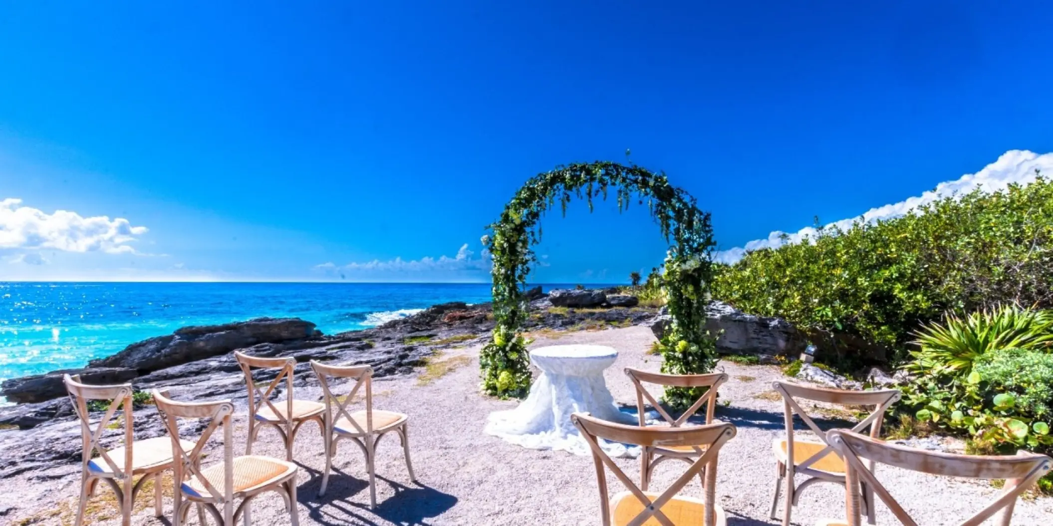 Rocky beachfront wedding ceremony with a floral arch and wooden chairs at Occidental at Xcaret, Playa Del Carmen