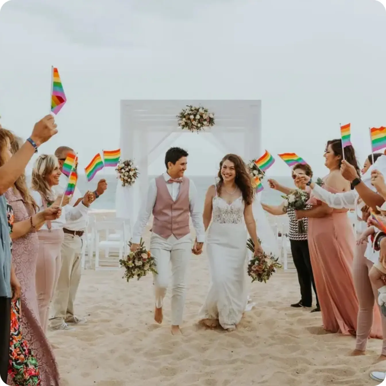 Newlyweds walking down the beach ceremony aisle through guests waving pride flags
