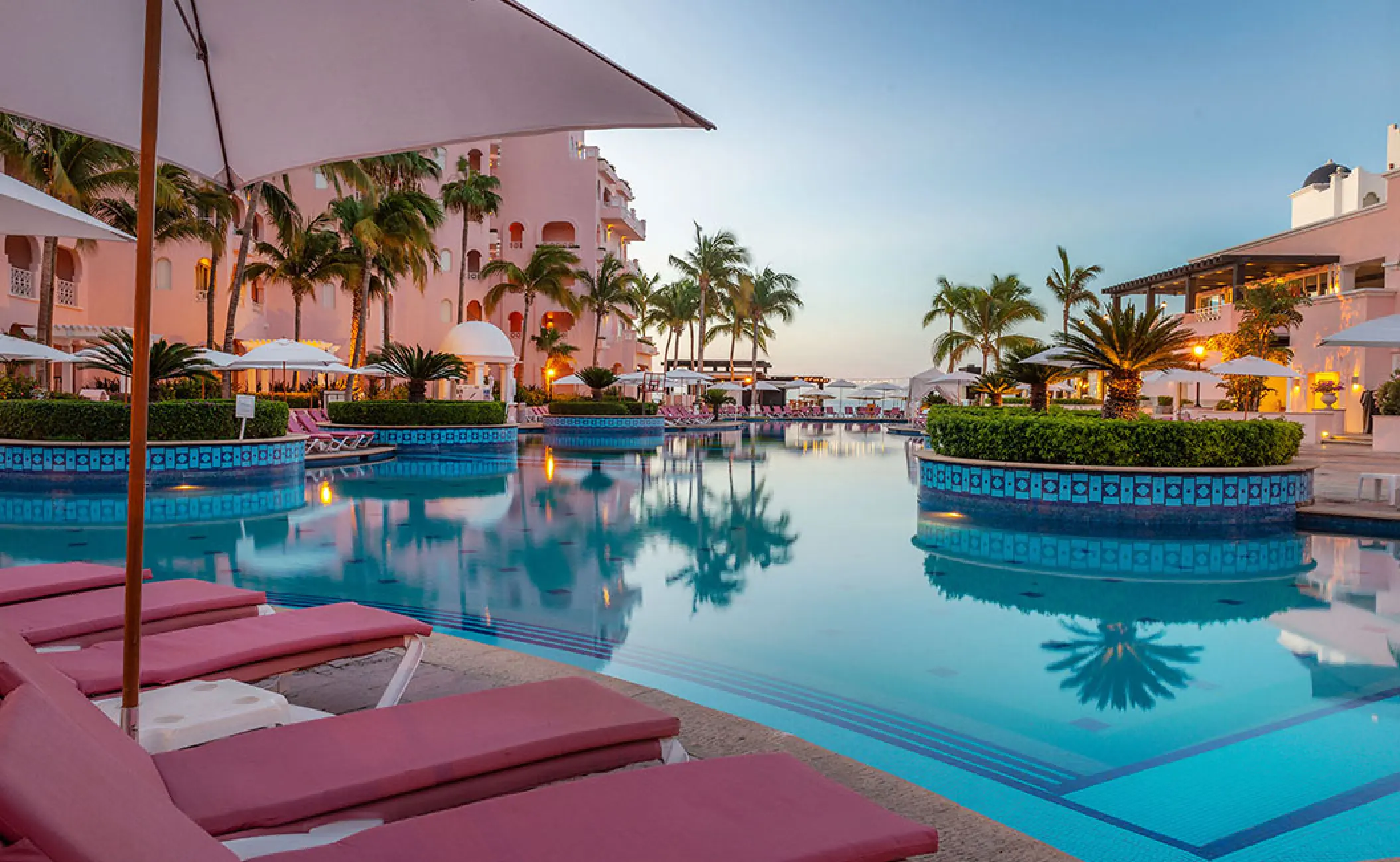 Pueblo Bonito Rose swimming pool with pink lounge chairs and palm trees at dusk, Cabo San Lucas