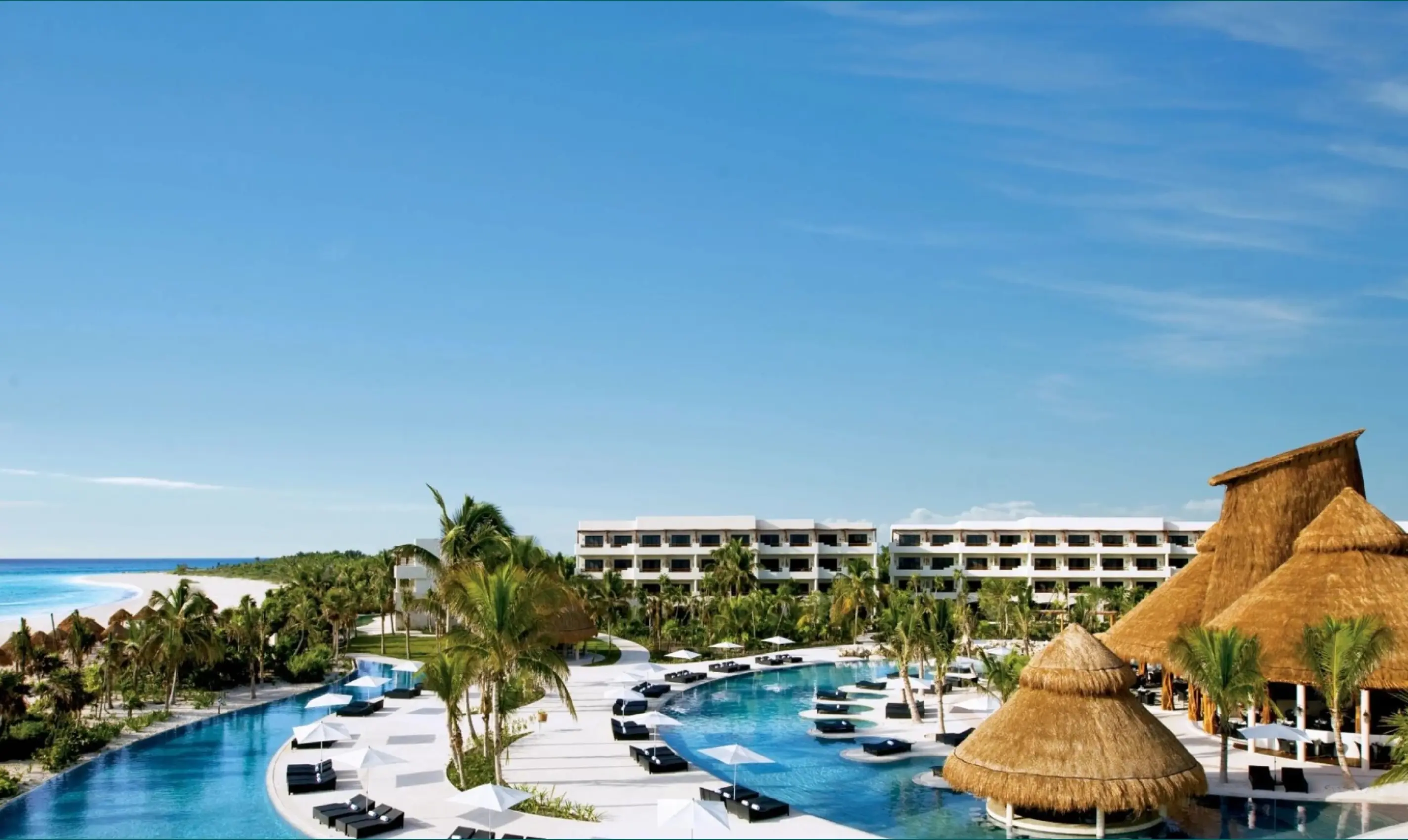Aerial view of a beachfront resort with thatched-roof palapas and winding pools overlooking the ocean