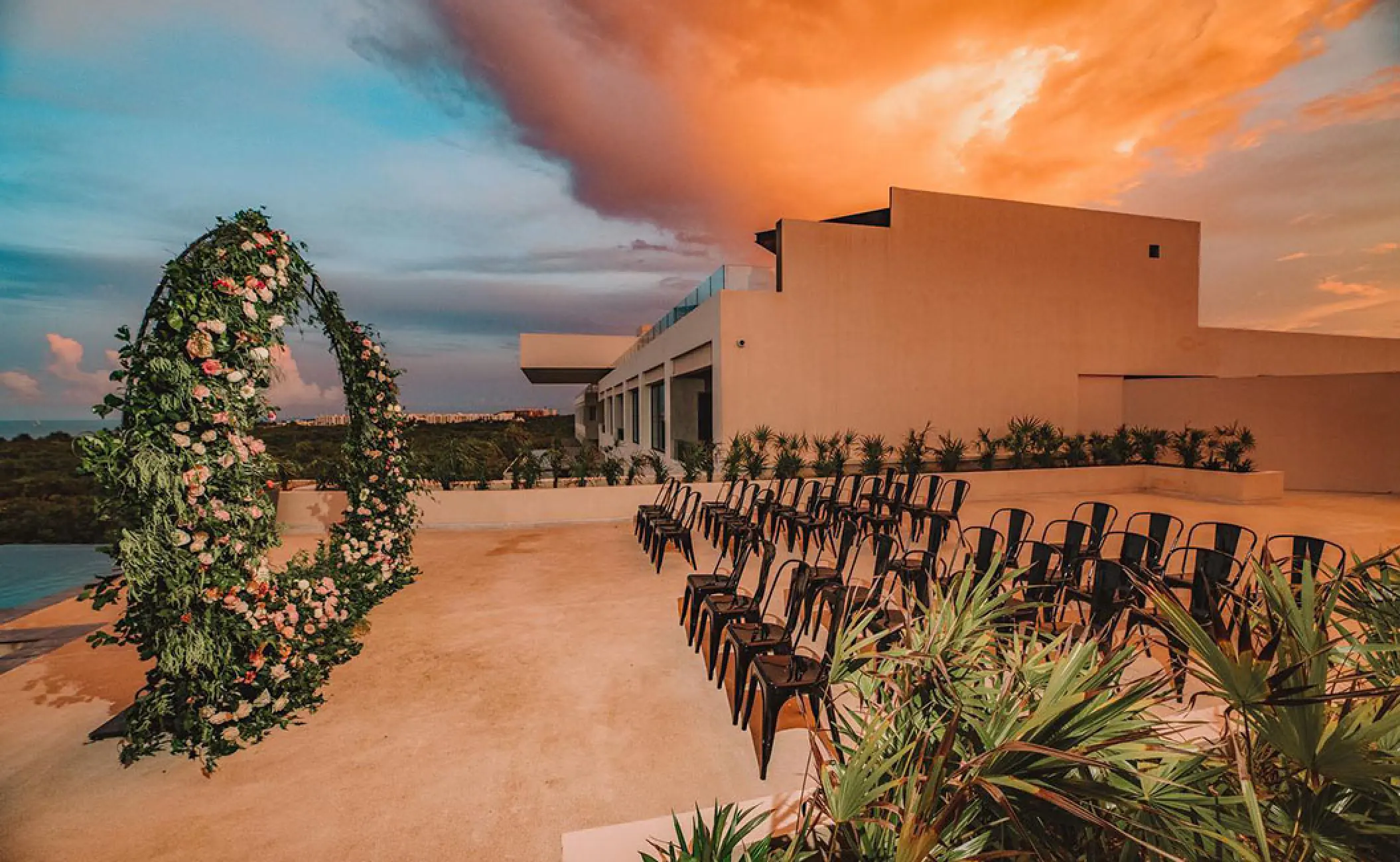 Terrace wedding ceremony with floral arch and black chairs at sunset, Atelier Playa Mujeres, Cancun