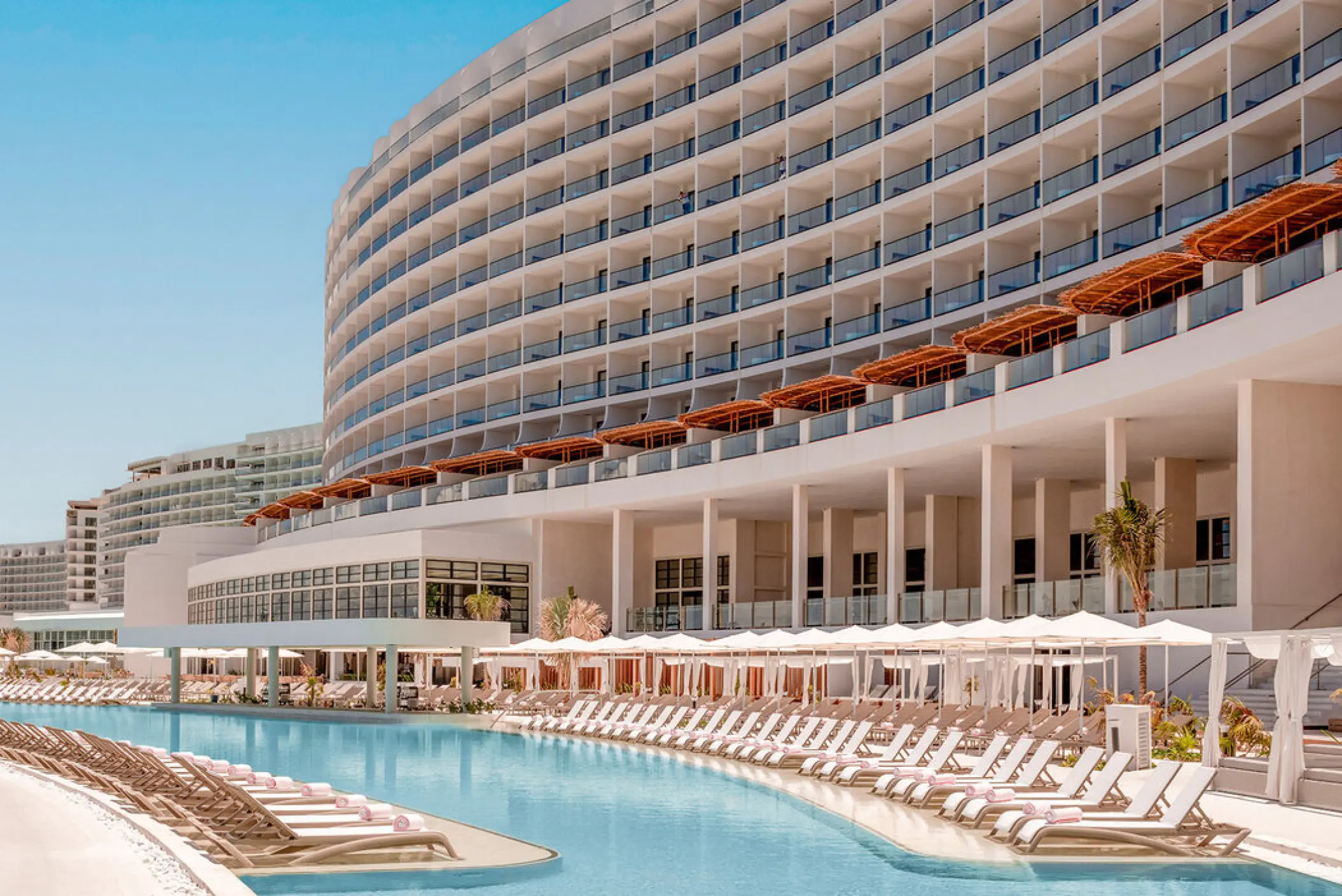 Curved hotel exterior and main swimming pool lined with lounge chairs at AVA Resort Cancun