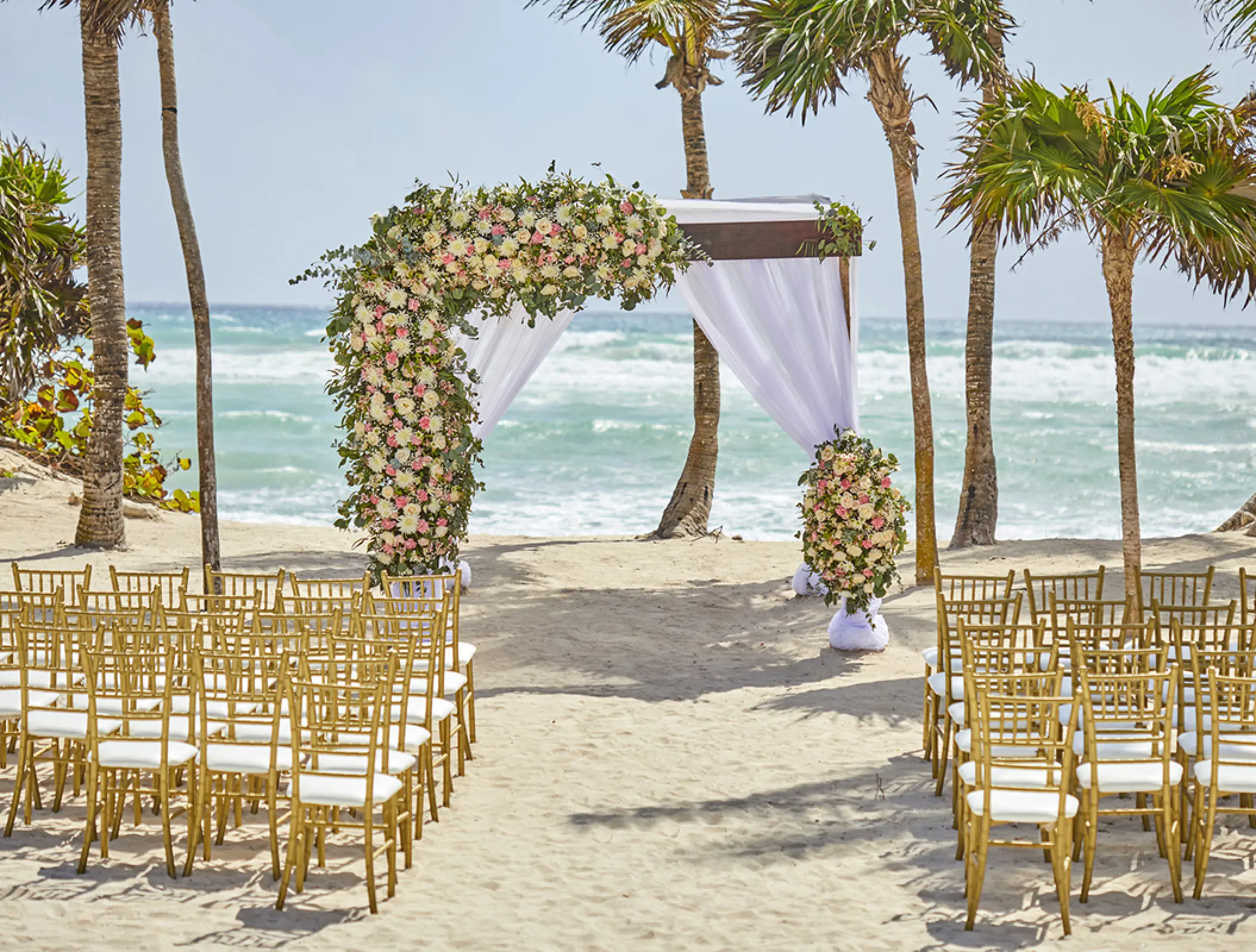 Beachfront wedding ceremony with a floral huppa and gold chairs at Bahia Principe Akumal, Tulum