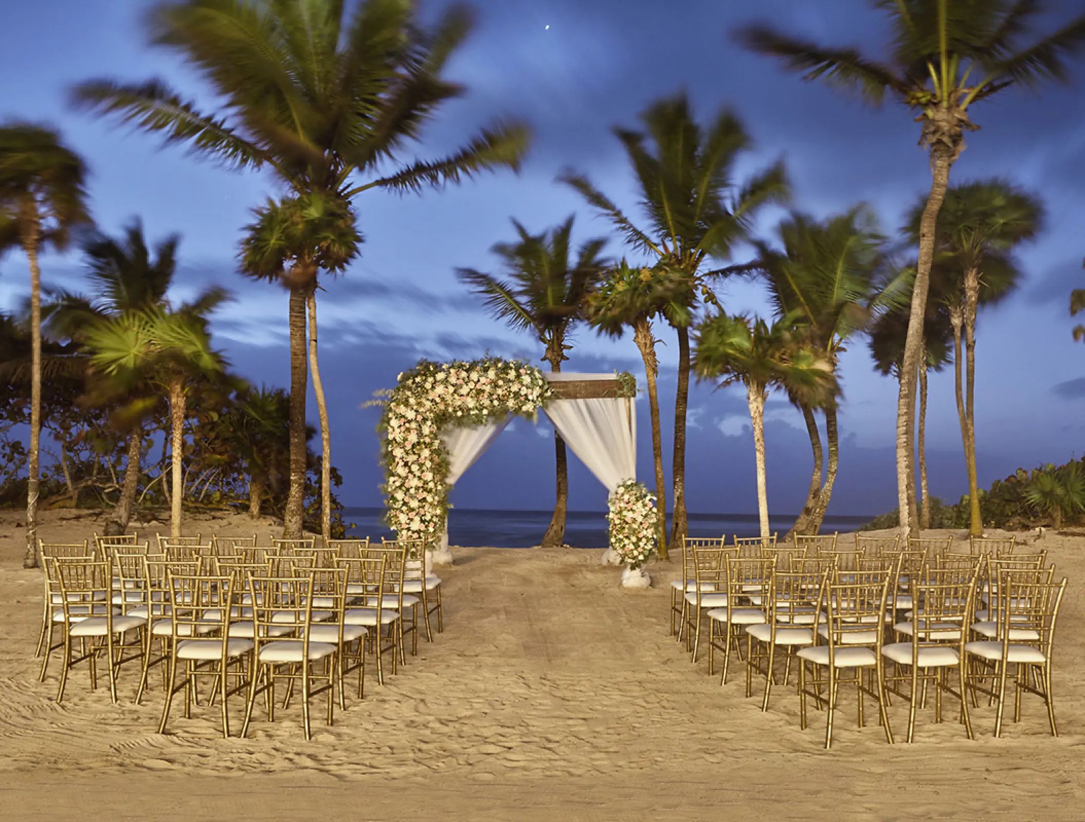 Beachfront wedding ceremony setup with white floral arch and gold chairs at Bahia Principe Akumal, Tulum