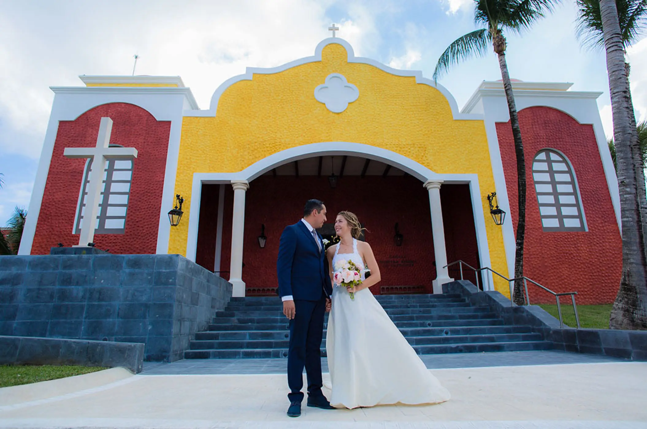 Bride and groom standing before the yellow and red Nuestra Senora de Guadalupe chapel at Bahia Principe Akumal, Tulum