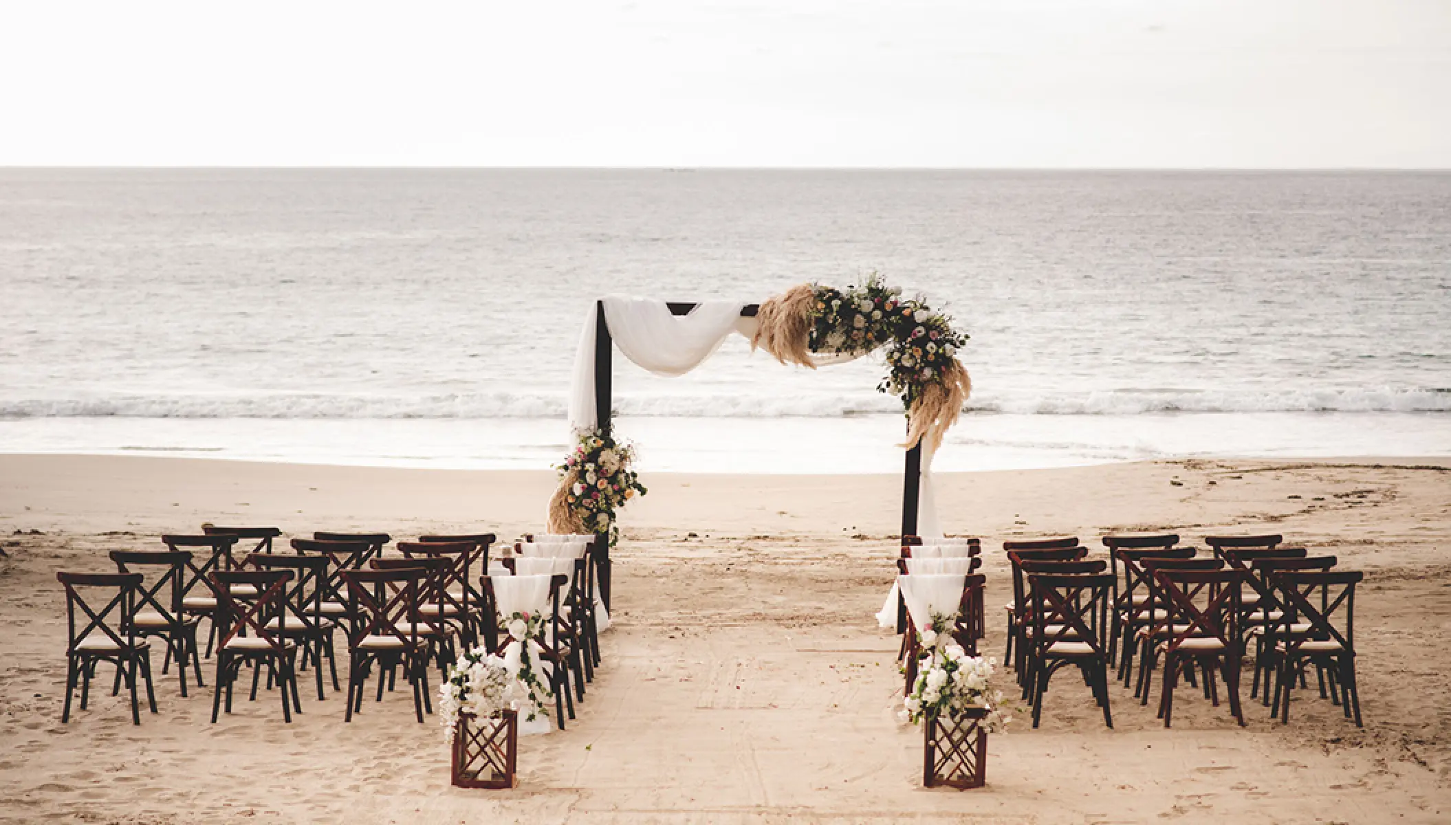 Beach wedding ceremony with a floral arch and wooden cross-back chairs at Conrad Punta de Mita, Puerto Vallarta