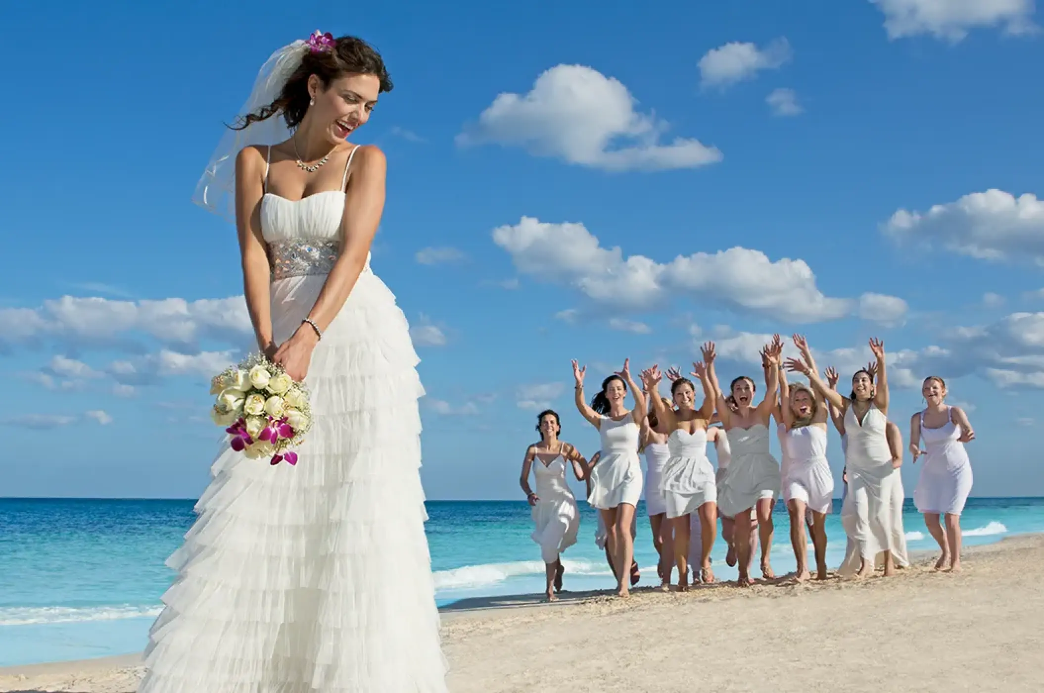 Bride holding a bouquet while bridesmaids run behind her on the beach at Dreams Cozumel, Cozumel