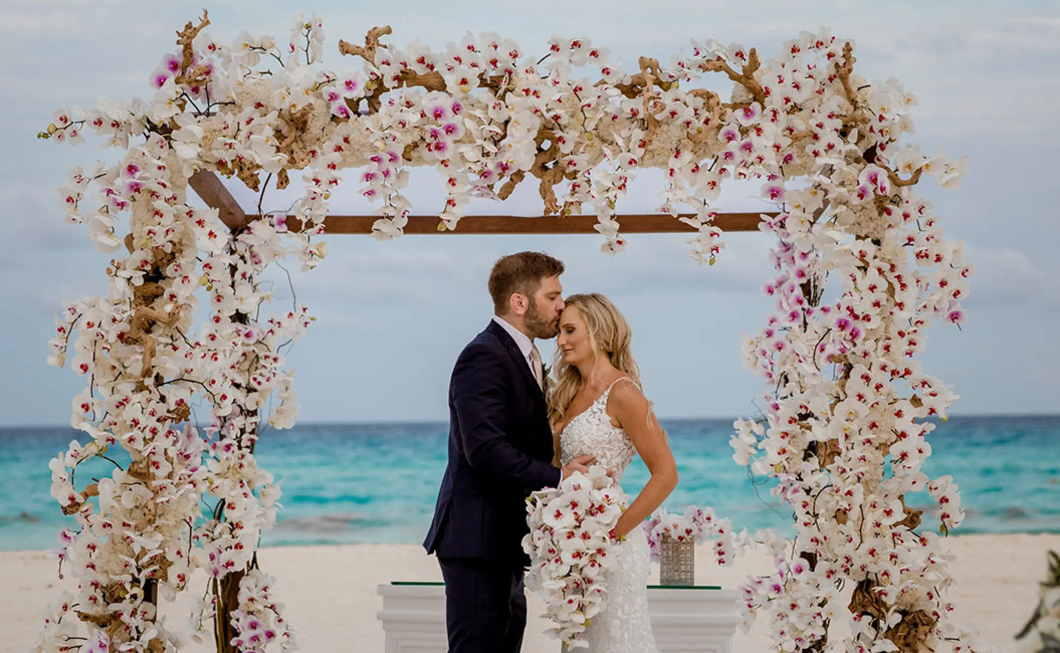 Groom kissing the bride's forehead under an orchid-covered arch at Dreams Cozumel Cape, Cozumel