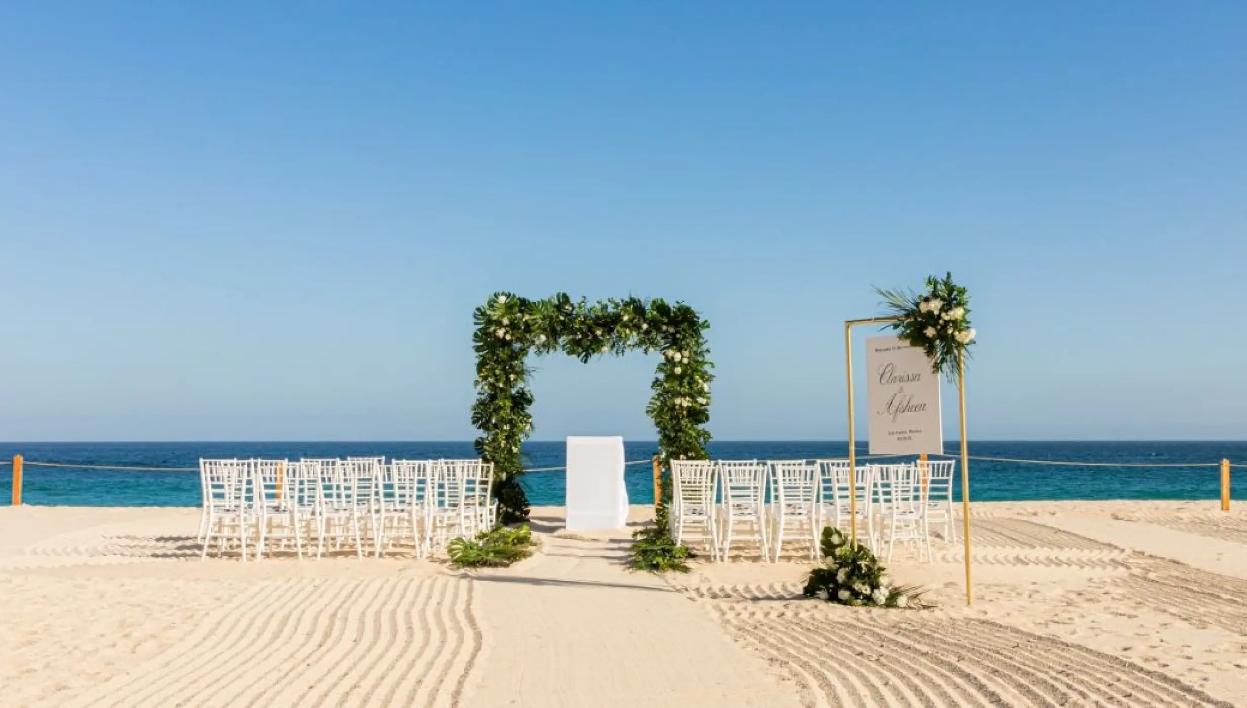 Beachfront wedding ceremony with greenery arch and white chairs at Dreams Los Cabos, Cabo San Lucas