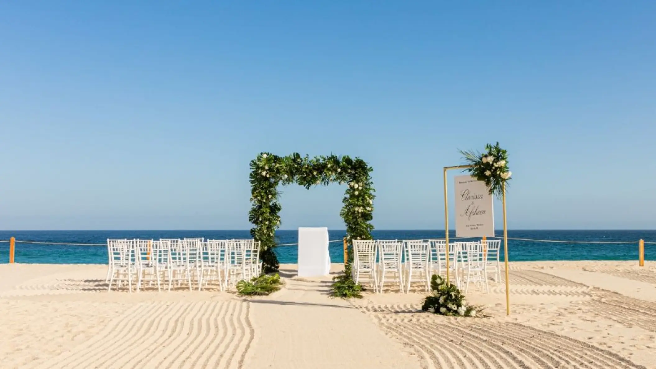 Beachfront wedding ceremony with greenery arch and white chairs at Dreams Los Cabos, Cabo San Lucas